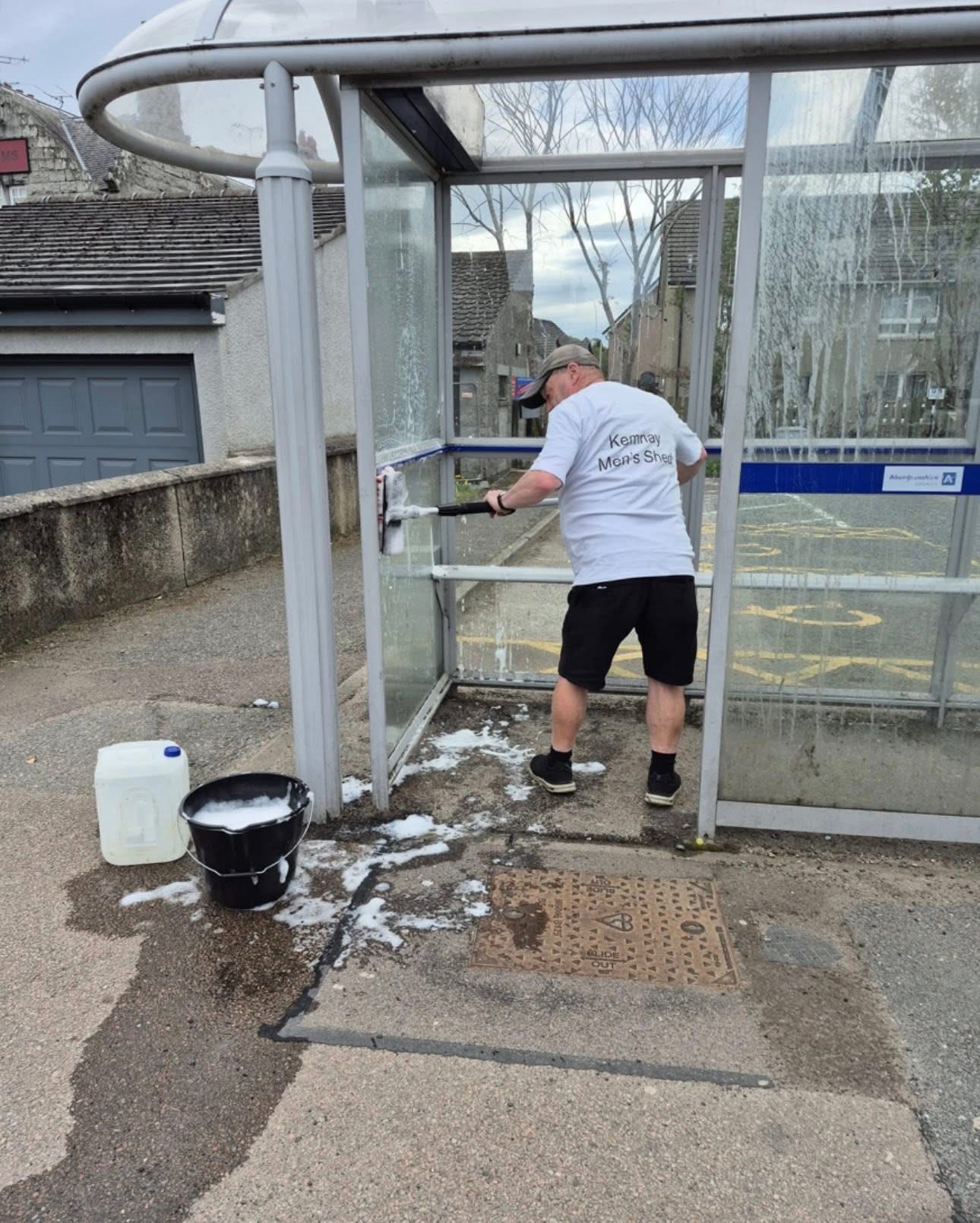 Member in a Kemnay Men's Shed t-shirt cleaning the village bus stop